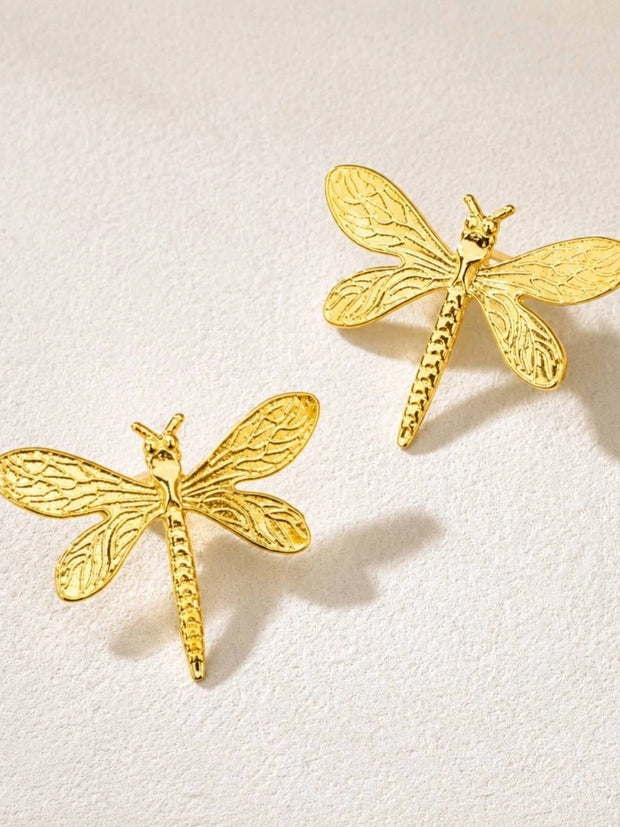 Two gold dragonfly stud earrings shown face-up on a white textured surface highlighting wing detail