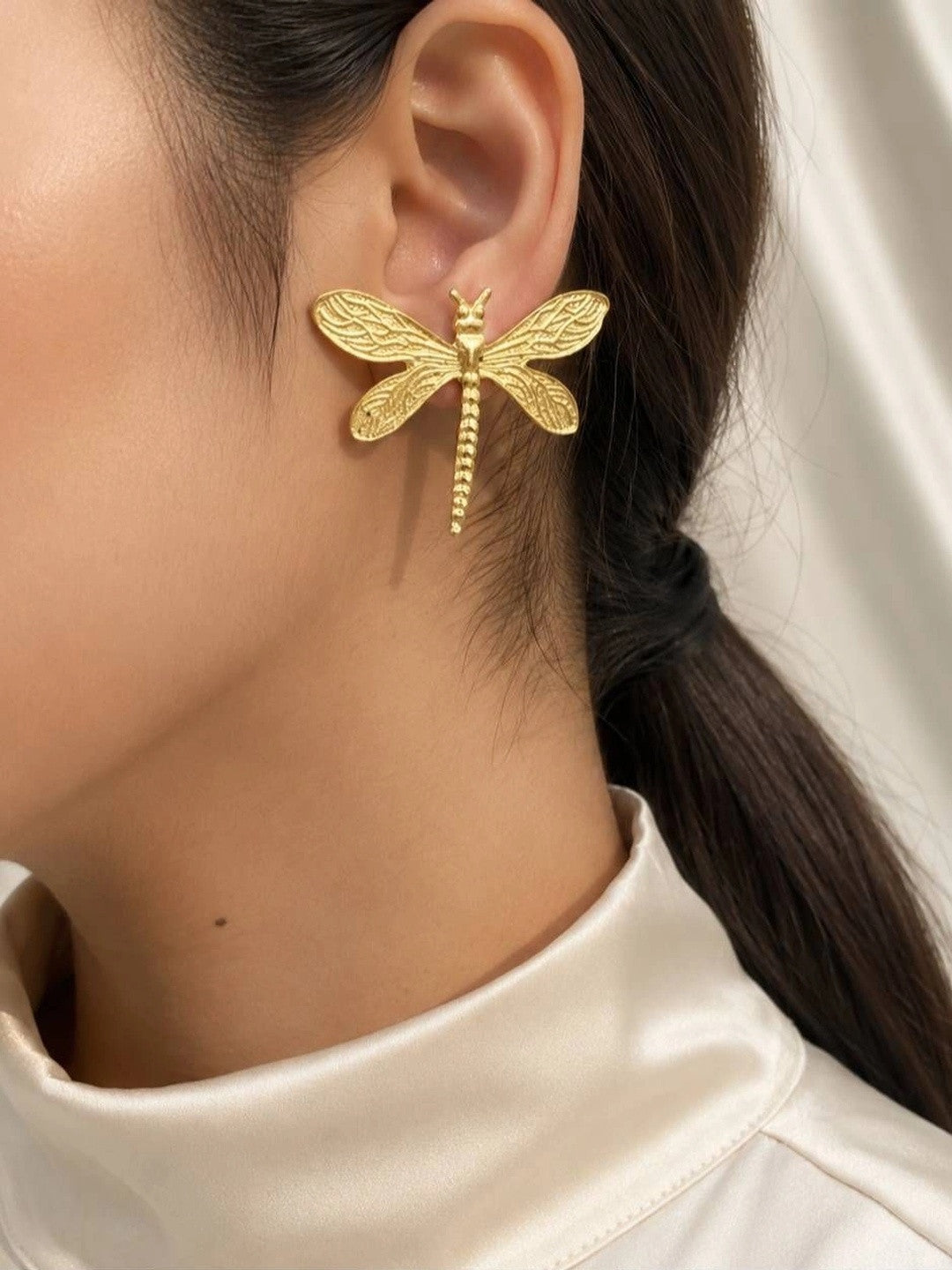 Gold dragonfly stud earring worn on a woman's ear against a white satin background