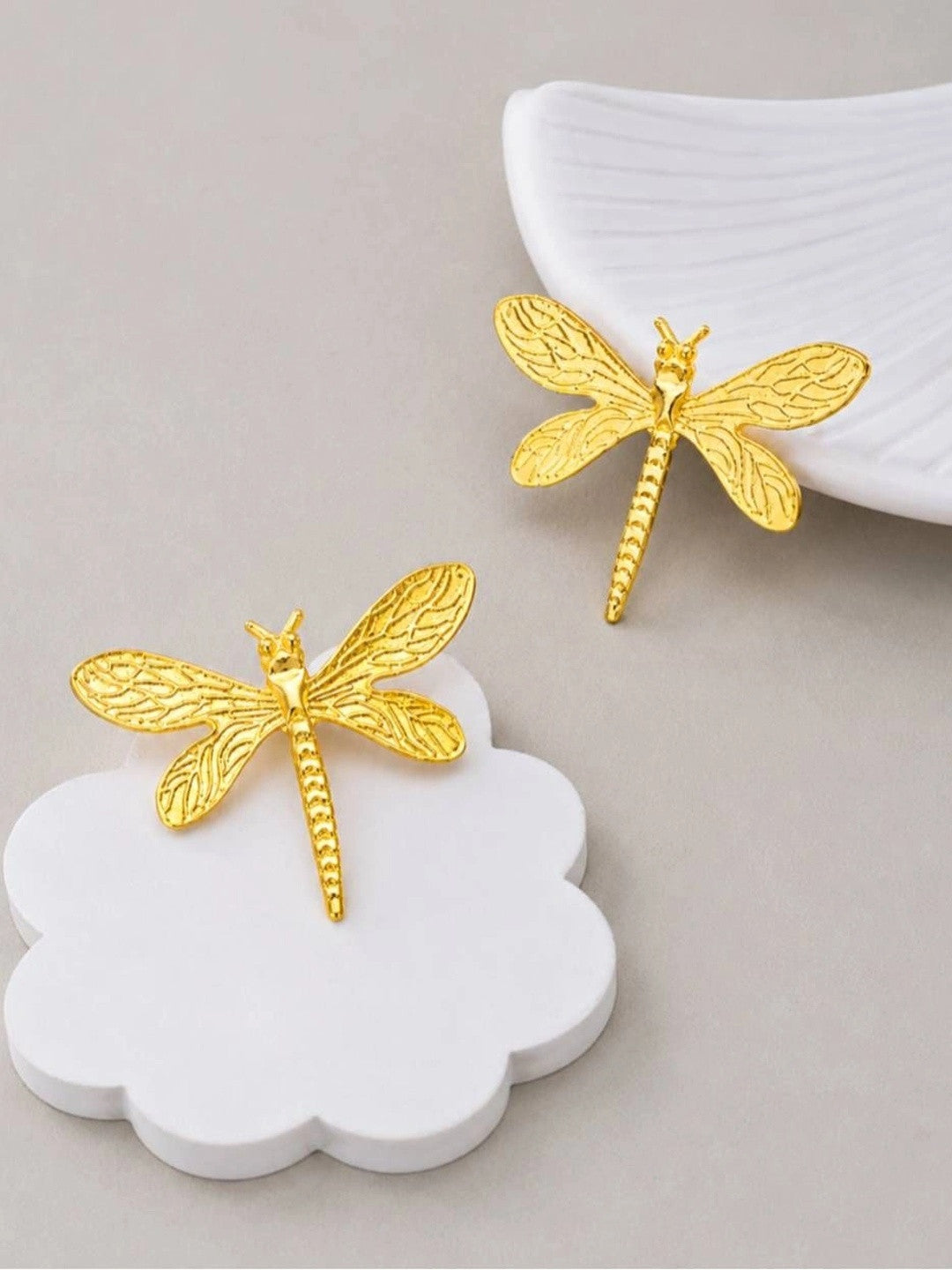 Gold-plated dragonfly stud earrings styled on white ceramic props against a grey background