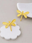 Gold-plated dragonfly stud earrings styled on white ceramic props against a grey background