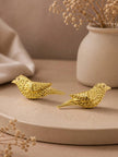 Two gold-plated sparrow stud earrings displayed on a beige stone surface with dried flowers and a ceramic vase