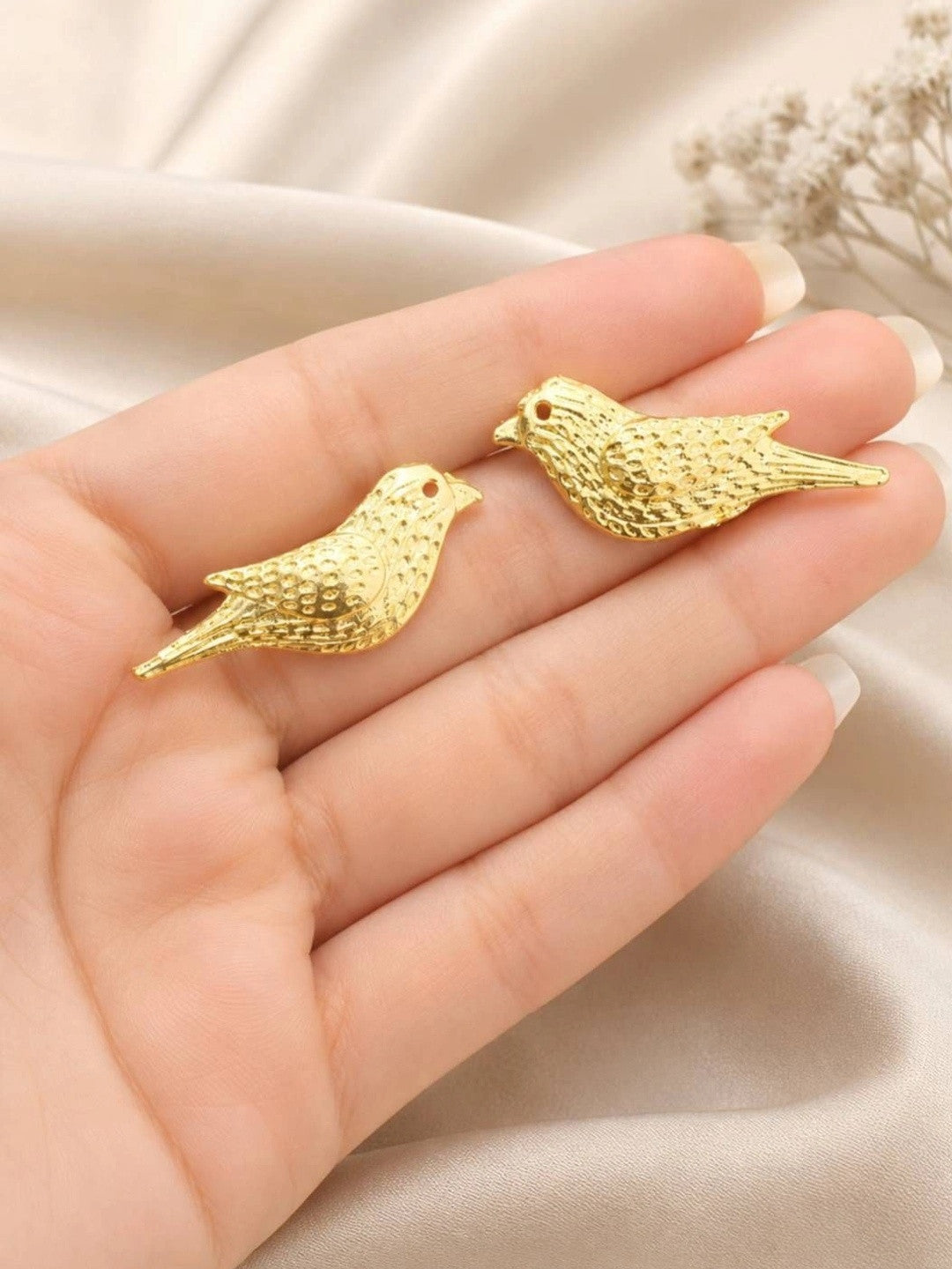 Pair of gold-plated bird stud earrings resting on an open palm showing scale and hammered feather texture