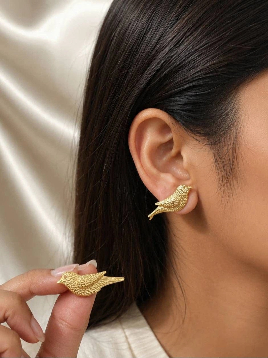 Gold bird stud earring worn on earlobe of a woman with dark hair, second earring held between fingers