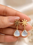 Close-up of gold-plated snowflake drop earrings held between fingers showing moonstone-like blue shimmer in teardrop