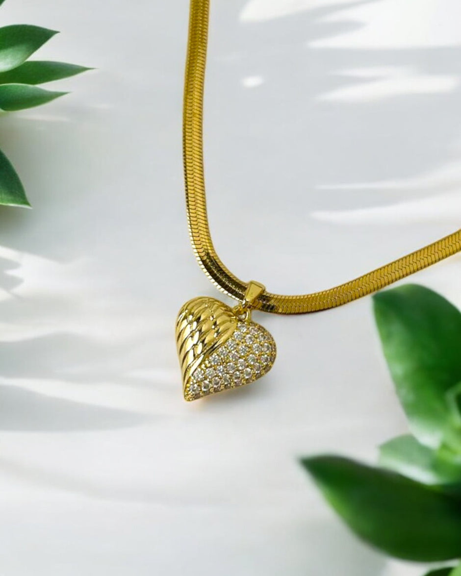 A gold heart-shaped pendant necklace with a textured and polished design, featuring a chain link and hanging over a white surface with green leaves in the background.