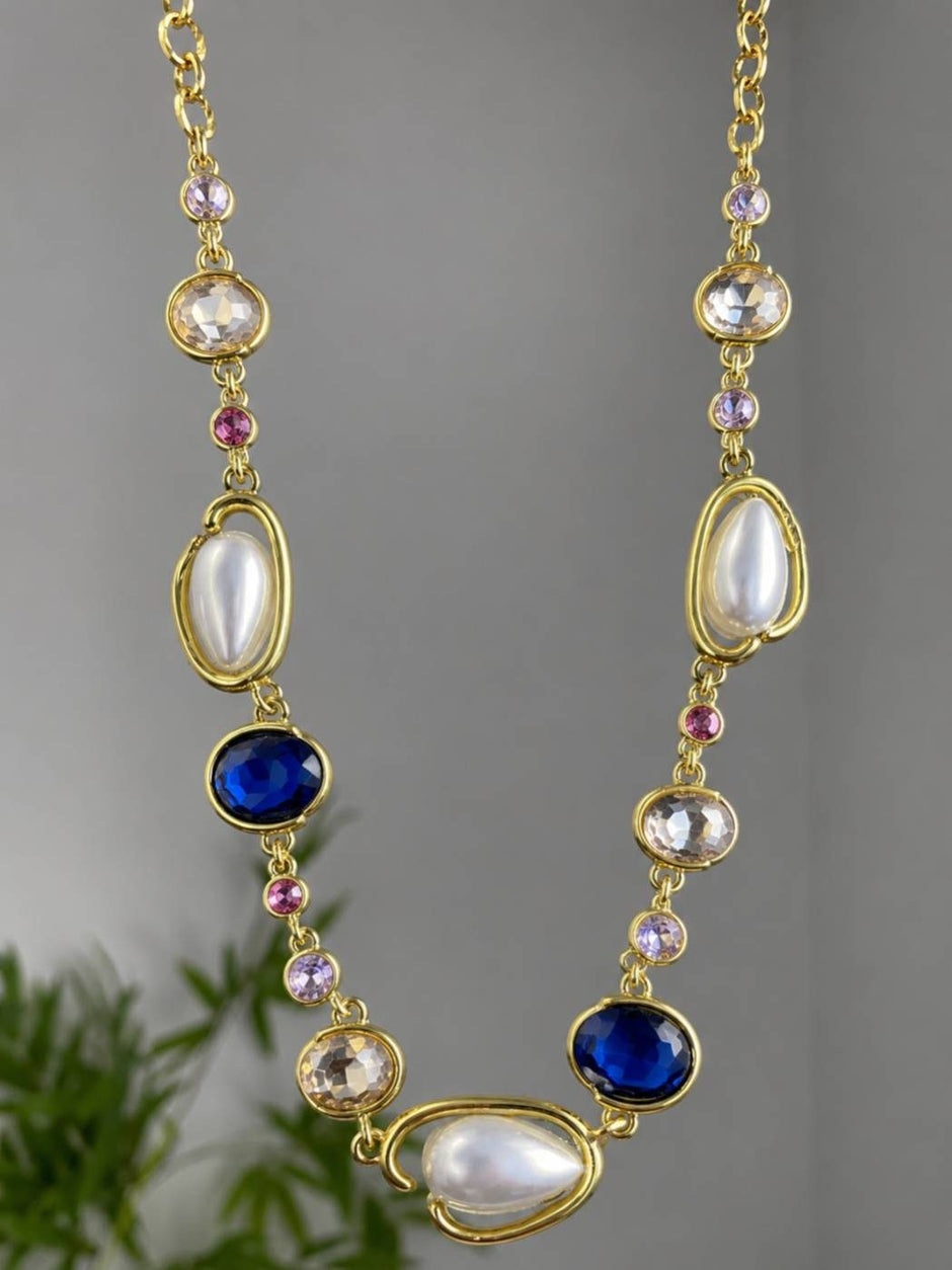 Gold-toned statement necklace with faux pearls and sapphire crystals hanging against a grey background