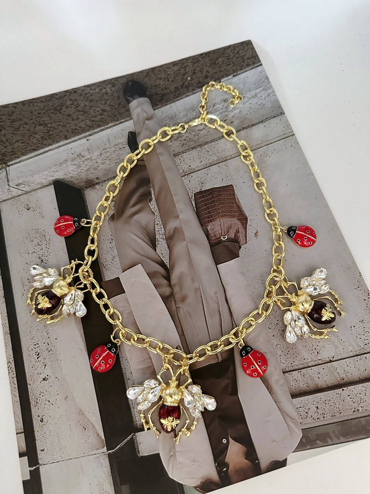 Gold-tone bee and ladybug necklace laid flat on a fashion magazine editorial background