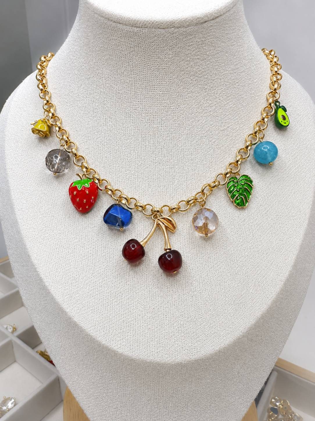 Gold-toned fruit charm necklace displayed on a white jewellery bust with enamel strawberry cherry and monstera leaf charms