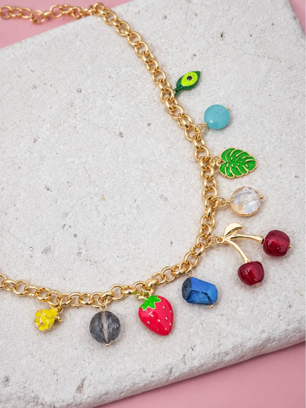 Close-up of gold rolo chain with enamel avocado monstera leaf blue bead strawberry and cherry charms on a stone surface