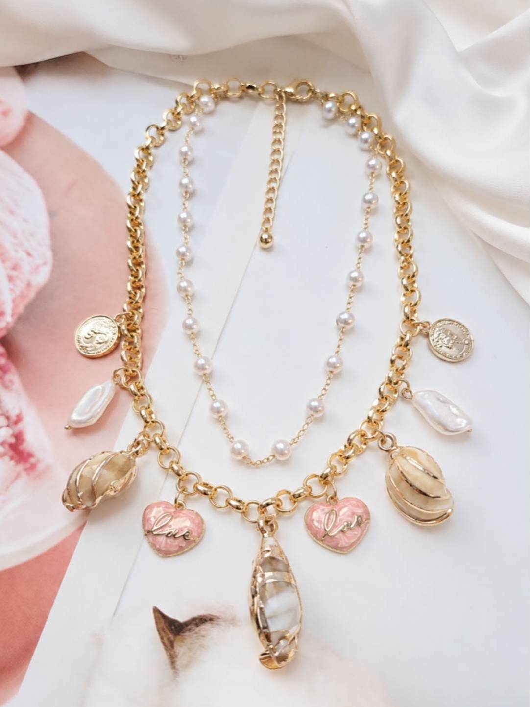 Flat lay of gold layered shell and pearl charm necklace on white surface beside a magazine and cat