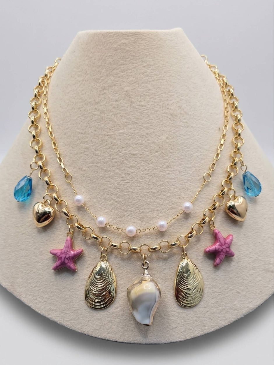 Gold-plated layered charm necklace with shell, starfish and crystal pendants displayed on a cream jewellery bust