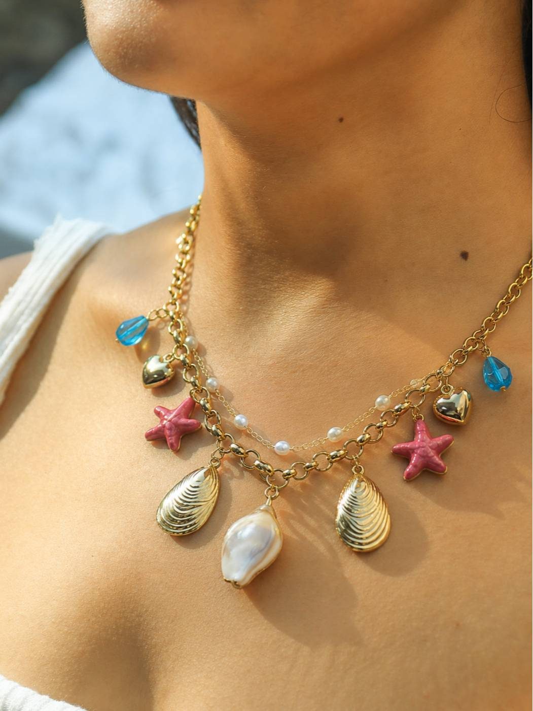 Woman wearing gold-plated layered shell and starfish charm necklace against a beach background