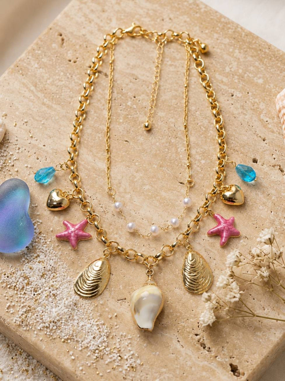Gold-plated ocean charm necklace laid flat on a sandy stone surface surrounded by seashells and dried flowers