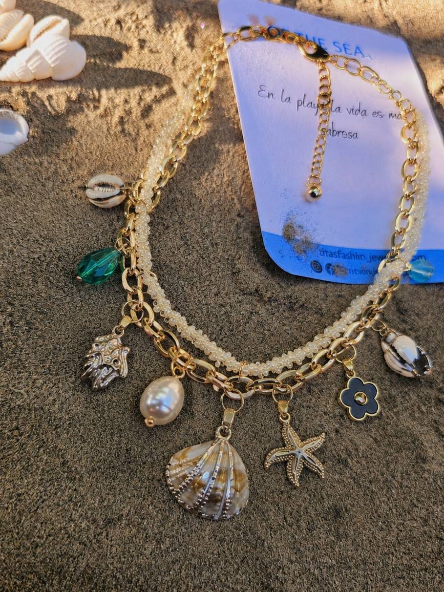 Gold-toned ocean charm necklace laid on sand next to branded card reading Of The Sea with Spanish tagline