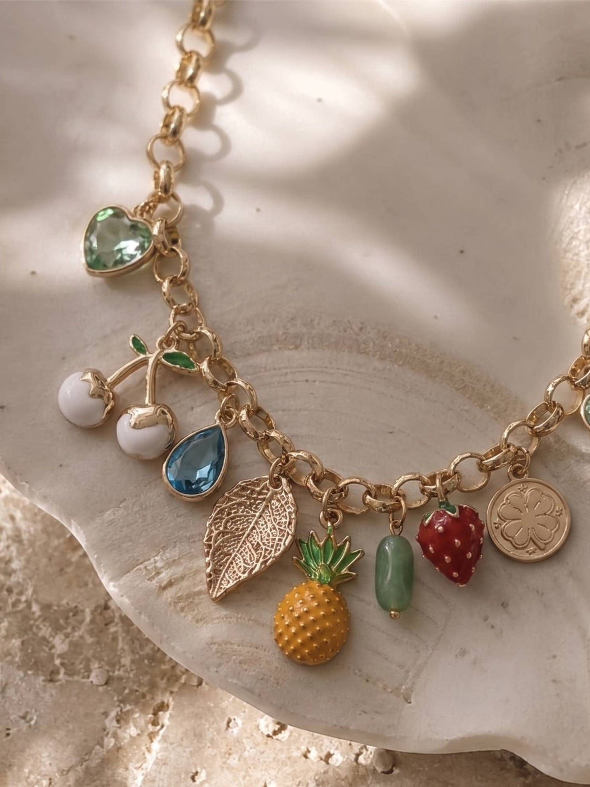 Close-up of fruit charm necklace on a stone plate highlighting pineapple, cherry, leaf and crystal teardrop charms in sunlight