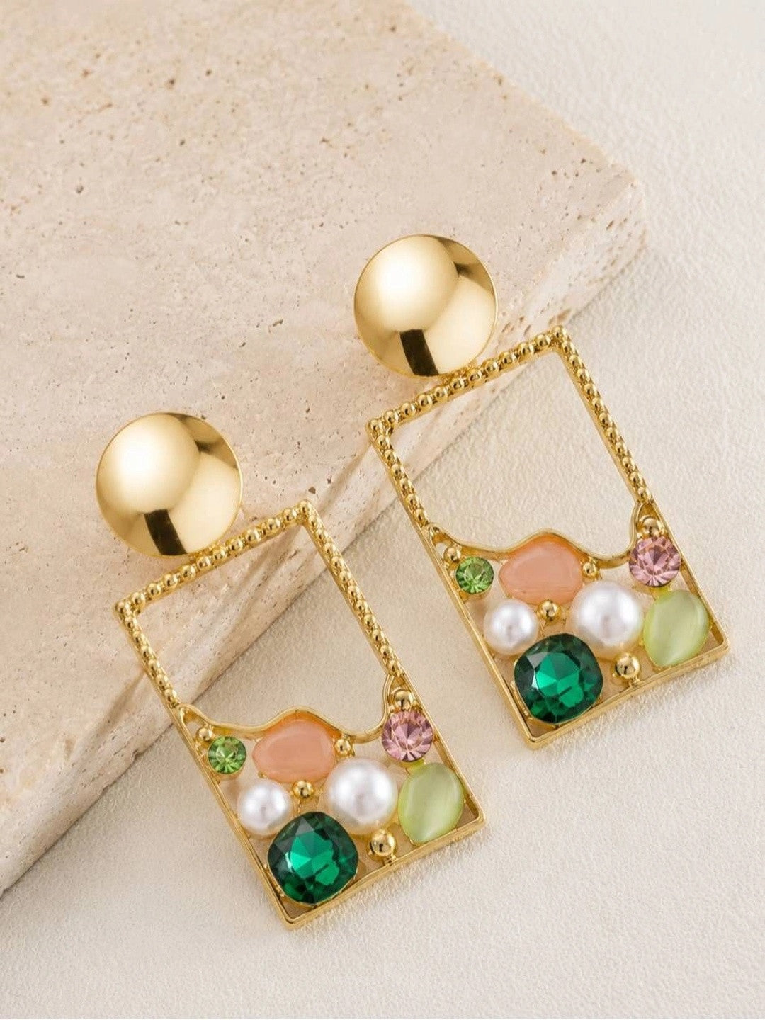 Pair of gold-plated rectangle drop earrings with green crystals, faux pearls and pink enamel on a beige stone surface