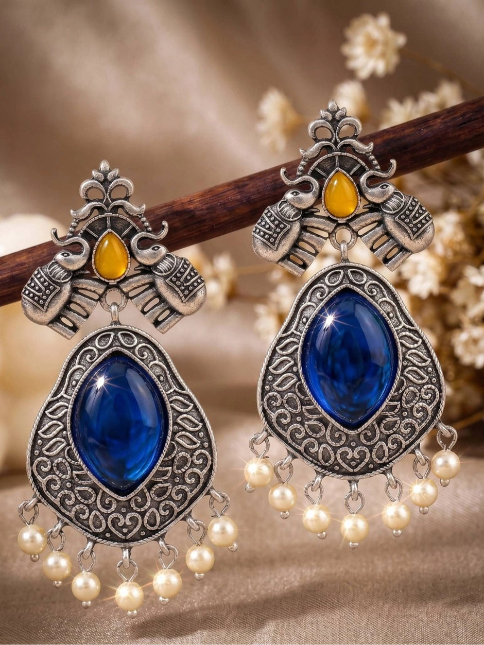 Pair of oxidised silver elephant chandbali earrings with blue cabochon and pearl drops propped on a wooden stick
