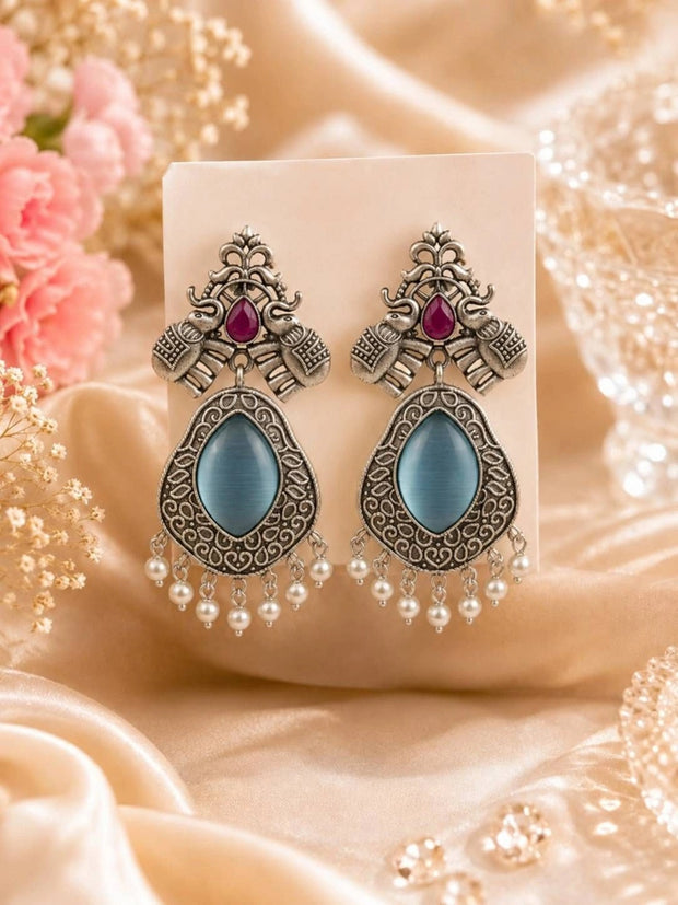 Pair of oxidised silver elephant teardrop earrings with blue cat-eye stone and pearl drops displayed on cream card against satin fabric