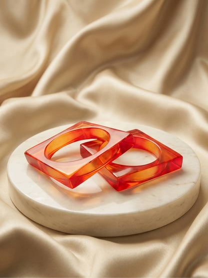 Two translucent orange resin square bangles resting on a white marble disc against cream satin fabric
