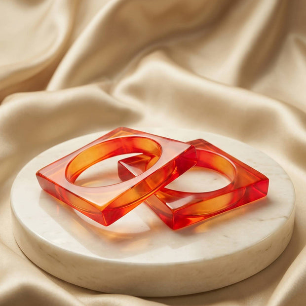 Two translucent orange resin square bangles resting on a white marble disc against cream satin fabric