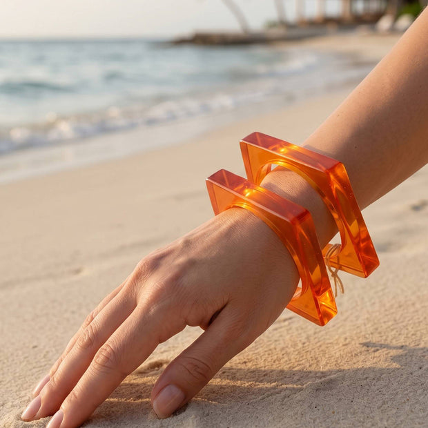 Two orange resin square bangles stacked on a woman's wrist resting on white sand at a tropical beach