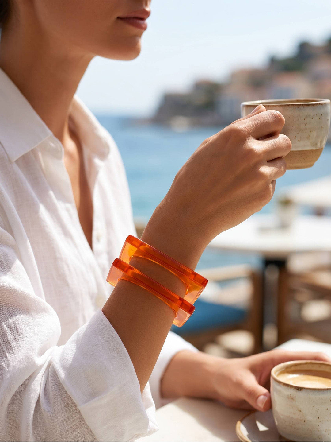 Woman in white linen shirt wearing stacked orange resin square bangles while holding a coffee cup at a seaside café