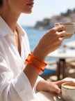 Woman in white linen shirt wearing stacked orange resin square bangles while holding a coffee cup at a seaside café