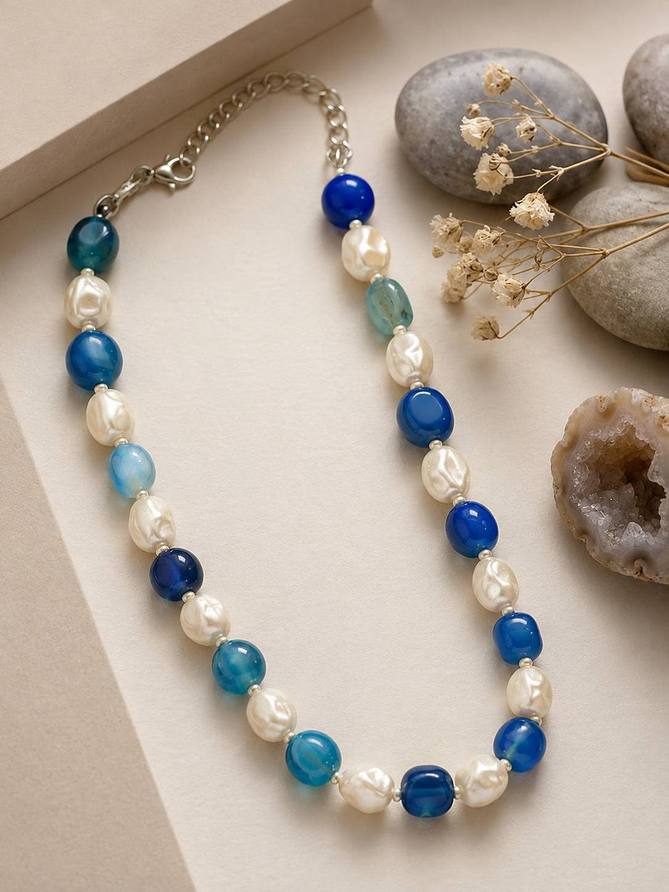 Blue agate and pearl beaded necklace laid on a white surface beside natural stones and a geode slice