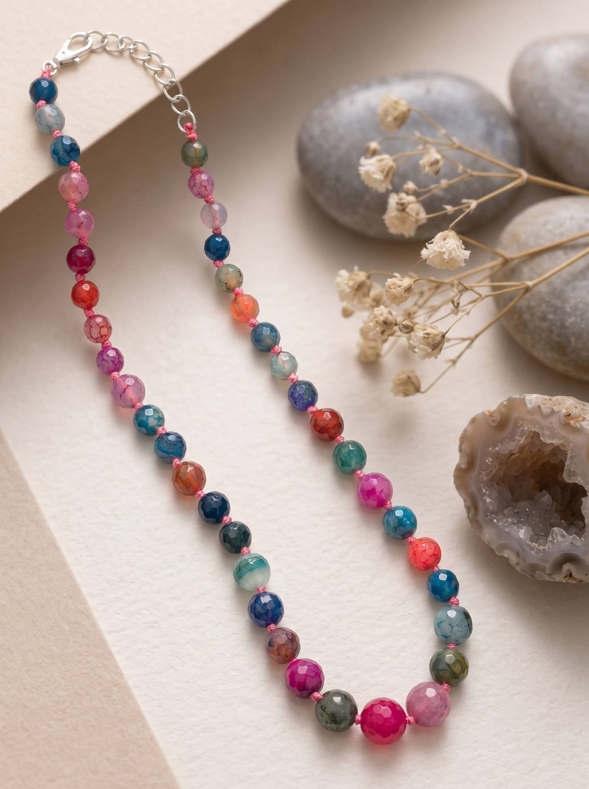 Multicolour agate bead necklace laid flat on white paper beside smooth grey stones and dried flowers