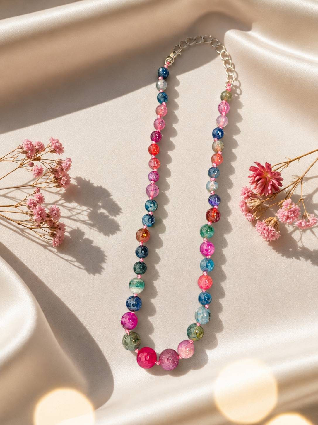 Colourful faceted agate bead necklace displayed on ivory satin fabric with pink dried florals and bokeh lights