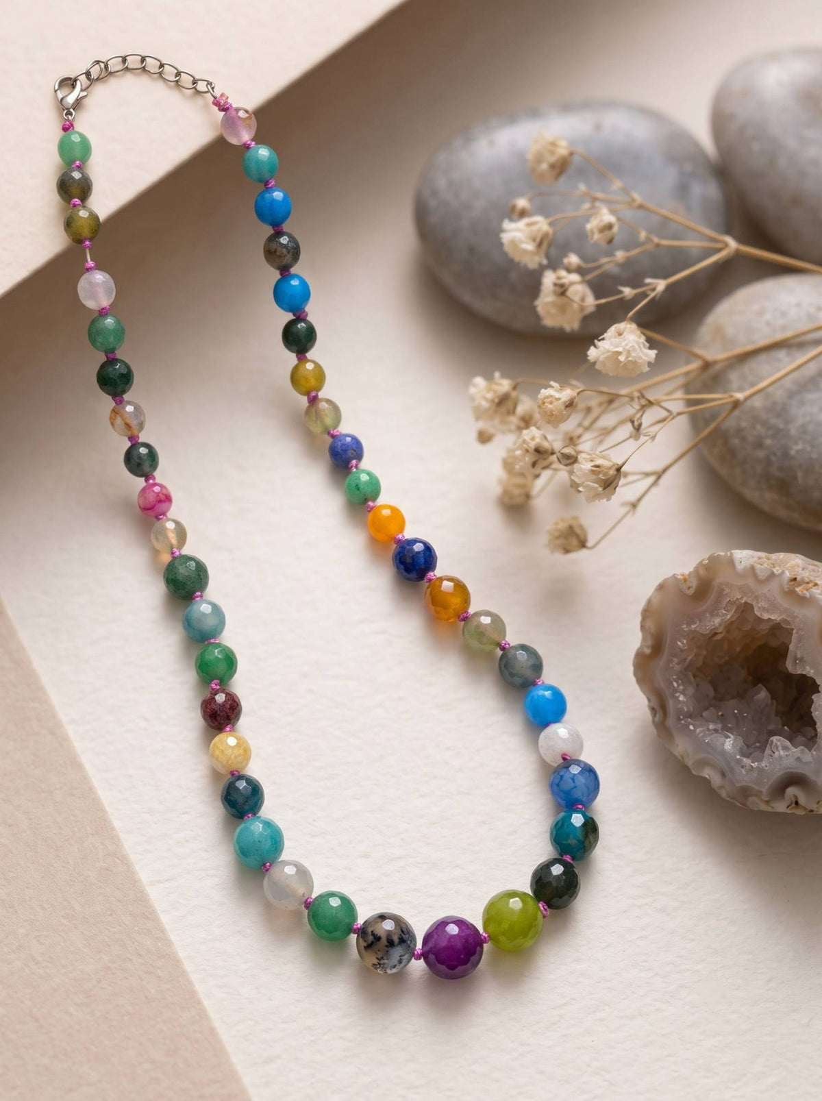 Multicolour stone bead necklace laid flat on white surface with smooth pebbles and dried flowers nearby