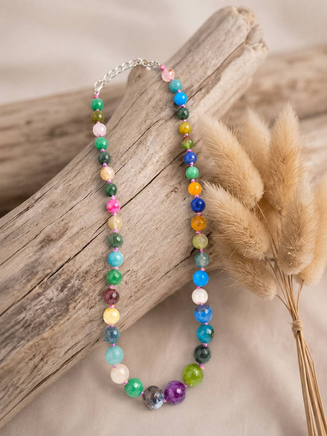 Colourful faceted bead necklace displayed on driftwood with dried pampas grass in warm natural light