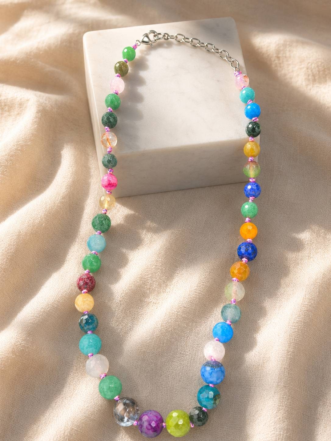 Rainbow stone bead necklace with silver lobster clasp draped over a white marble block on cream fabric