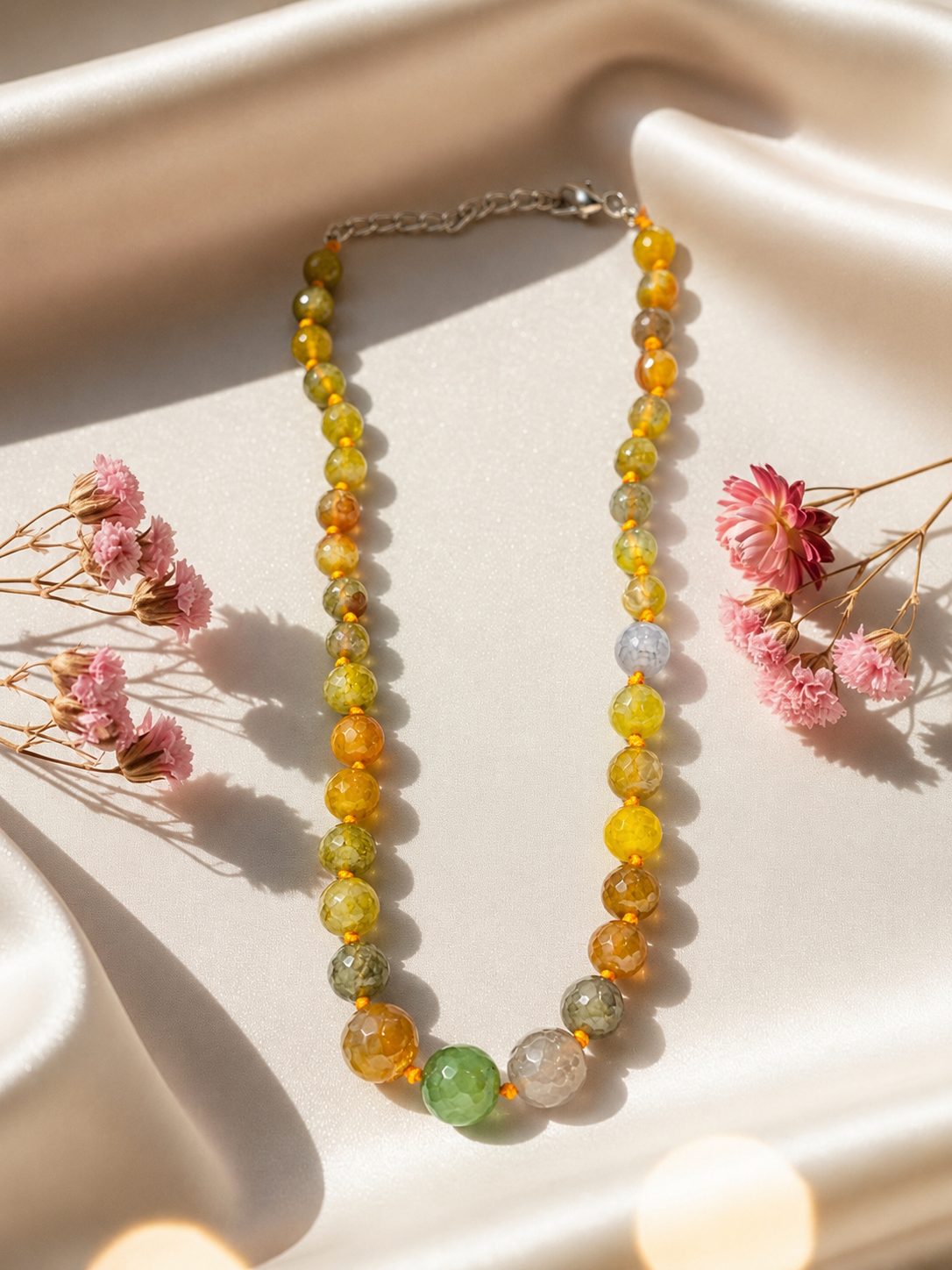 Multicolour faceted agate bead necklace laid flat on ivory satin with pink dried flowers
