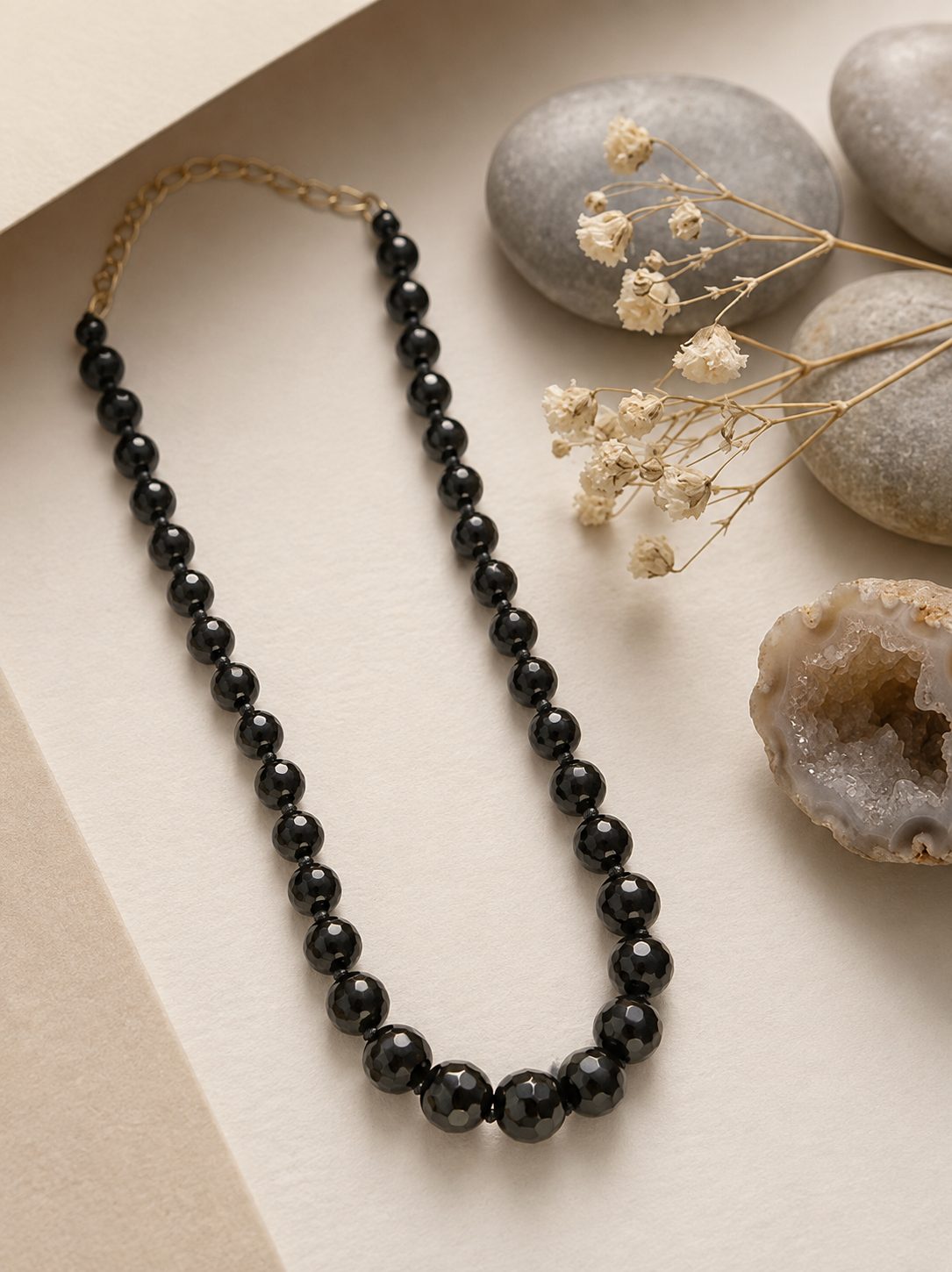 Black graduated faceted bead necklace laid flat on cream paper beside smooth grey pebbles and dried white flowers