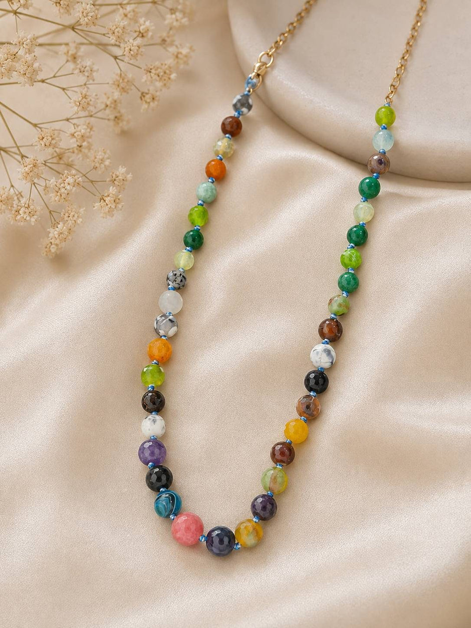 Colourful mixed stone bead necklace with blue seed spacers displayed on cream satin fabric with gold chain clasp