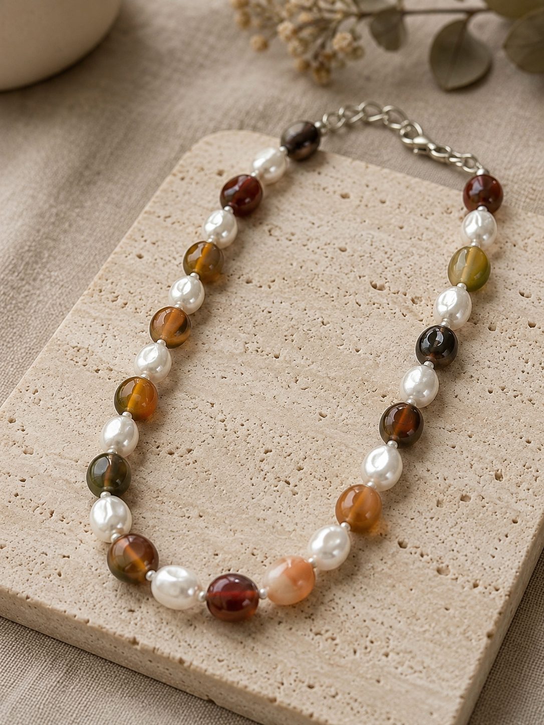 Agate and baroque pearl beaded choker necklace laid flat on a textured travertine tile with dried botanicals in the background