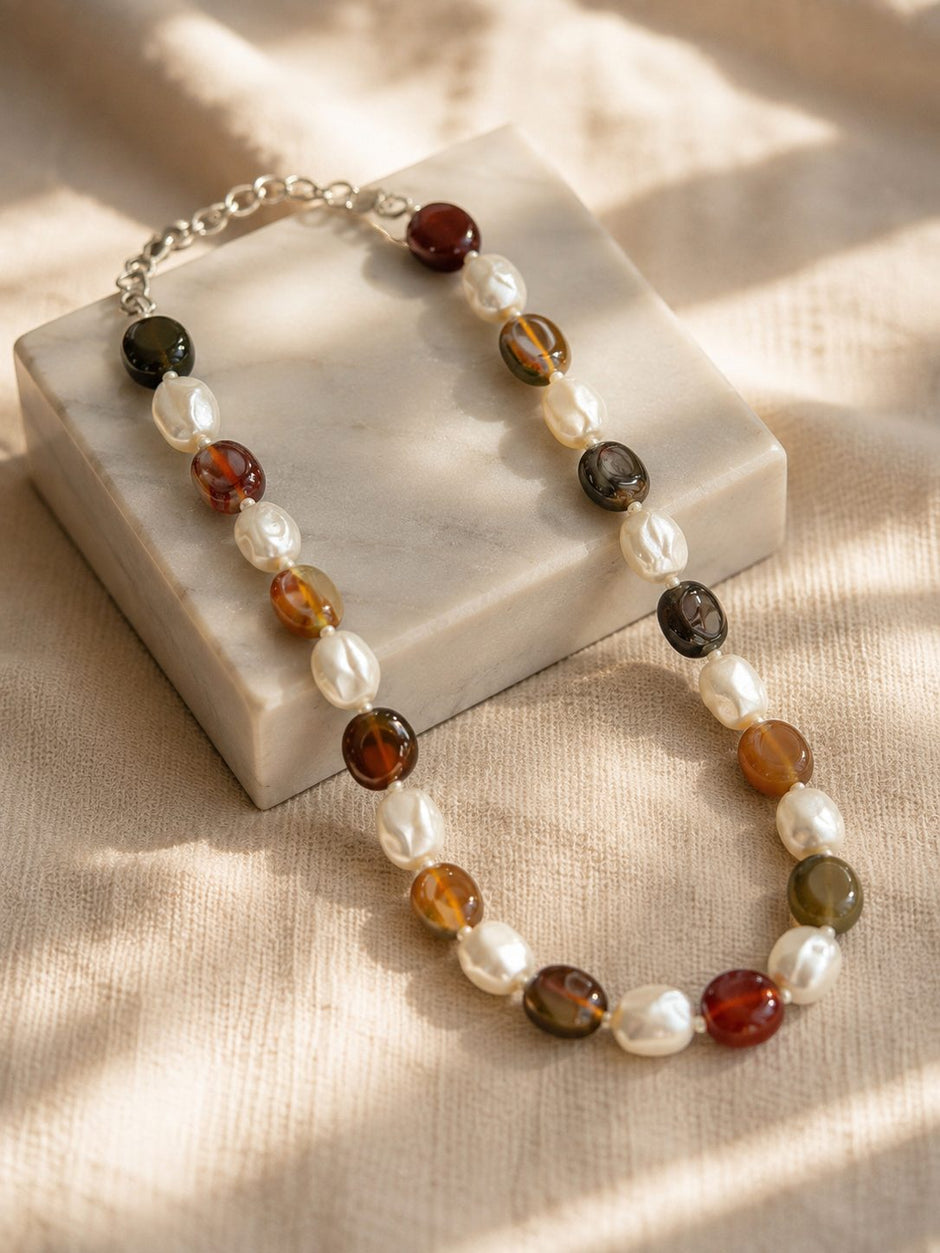 Earthy brown and ivory beaded choker necklace draped over a white marble block with soft natural light and linen fabric background