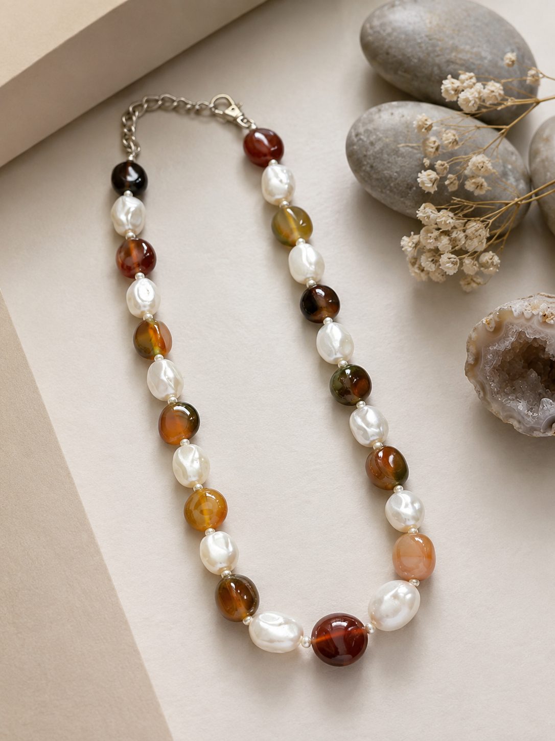 Full-length beaded choker necklace laid diagonally on a cream surface beside smooth river stones, dried flowers and a raw agate slice