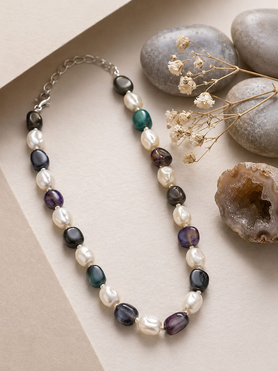 Pearl and tumbled stone necklace displayed on a light paper tray beside smooth pebbles and a geode slice