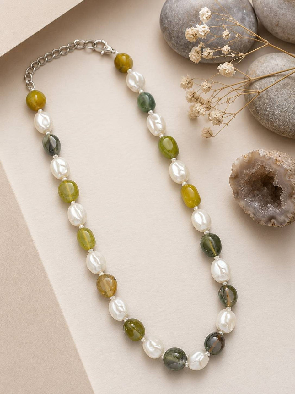 Green agate and white baroque pearl beaded necklace laid flat on beige surface with pebbles and dried flowers