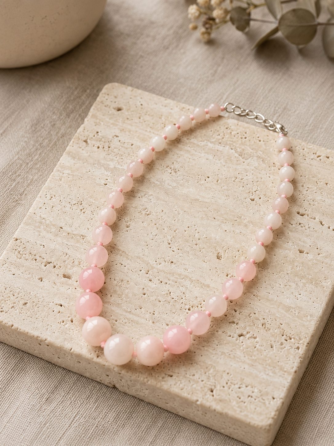 Full view of pink quartz bead necklace laid on a travertine tile with eucalyptus leaves in the background