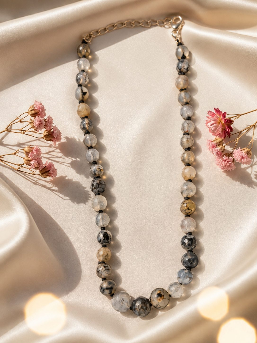 Grey marbled bead necklace arranged in a U-shape on ivory satin fabric beside pink dried florals