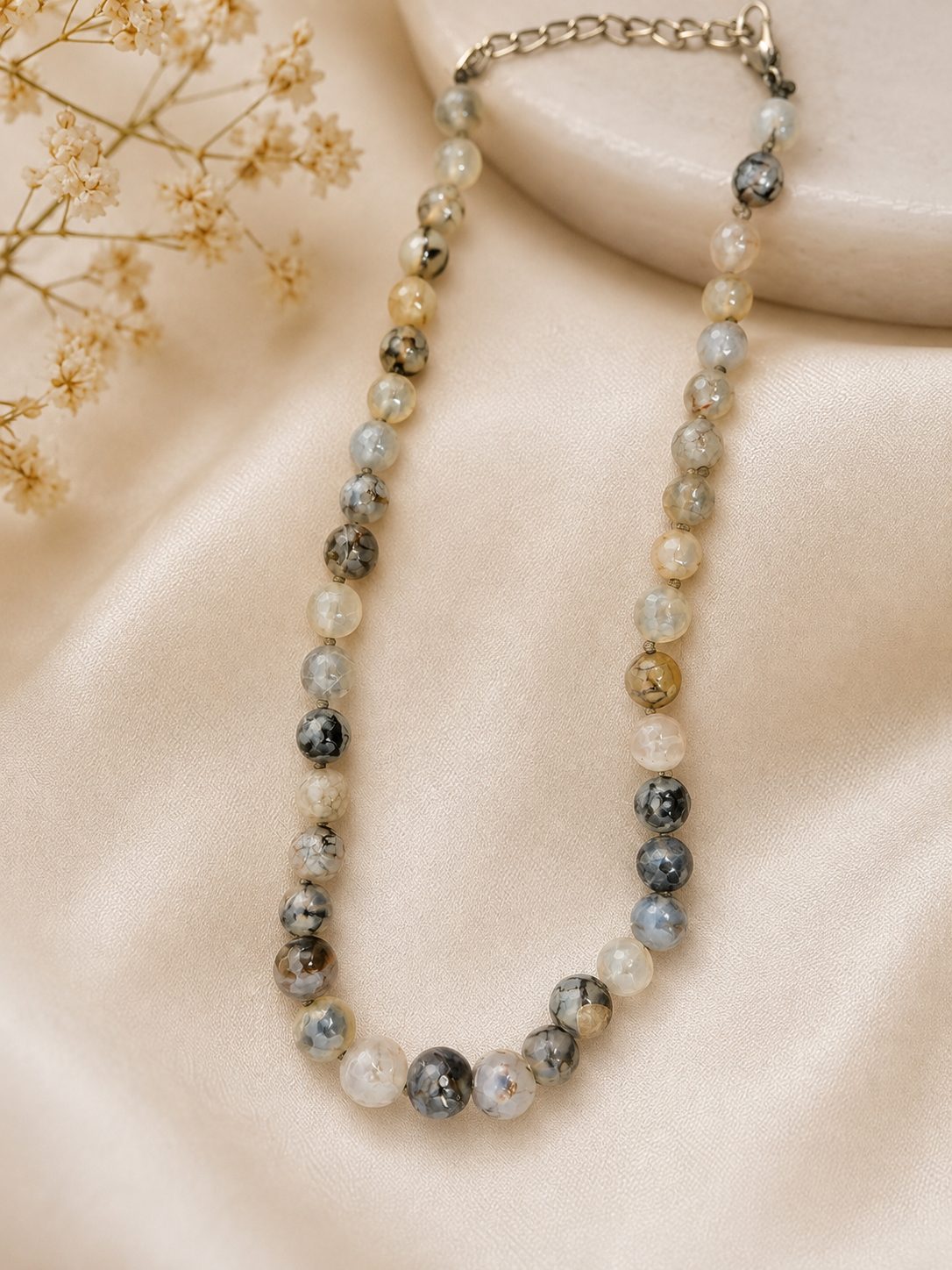 Grey agate-style graduated bead necklace with silver extension chain laid on cream satin with gypsophila