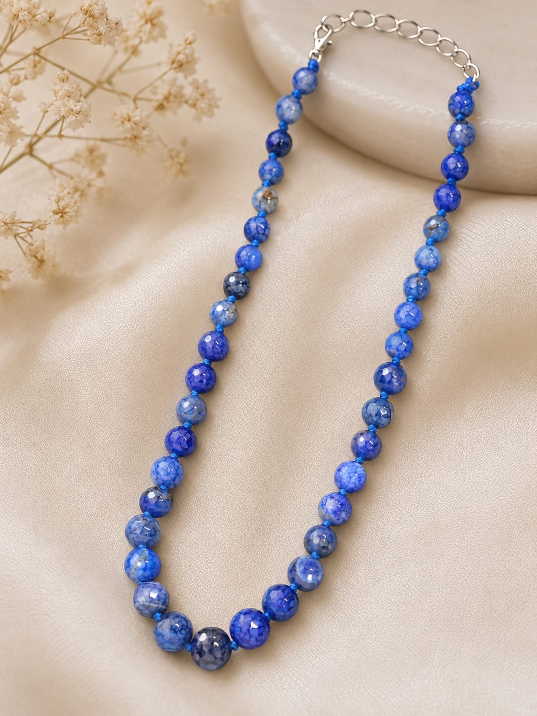 Blue crackle agate bead necklace laid flat on a cream satin surface with dried flowers