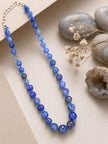 Blue faceted agate bead necklace displayed on a white surface beside smooth grey pebbles and a geode
