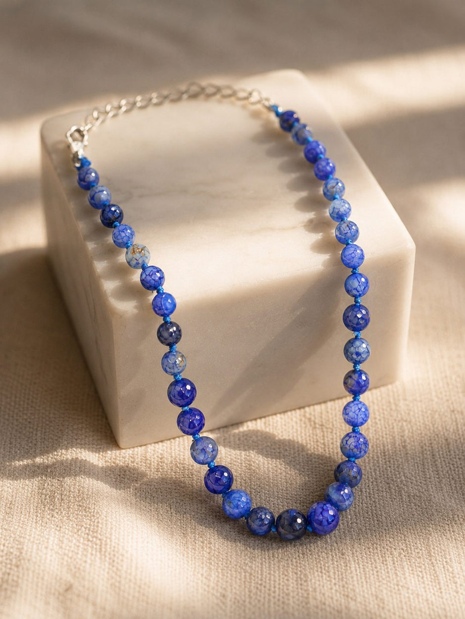 Blue crackle agate bead necklace draped over a white marble block in natural sunlight