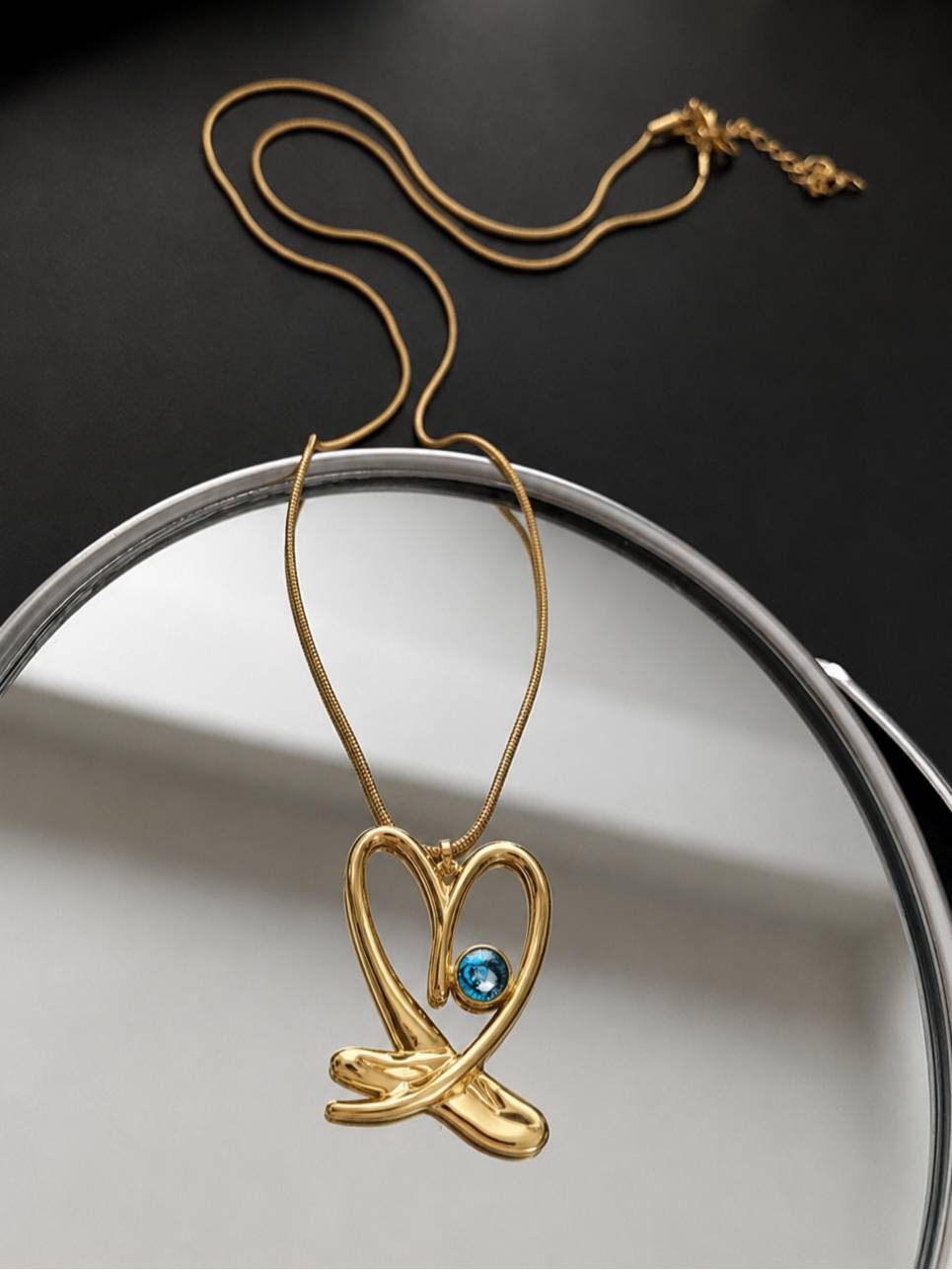 Gold evil eye heart pendant necklace draped over the edge of a round mirror on a white surface