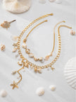 Both gold-tone shell necklaces styled together on a sandy white surface surrounded by real seashells and coral pieces