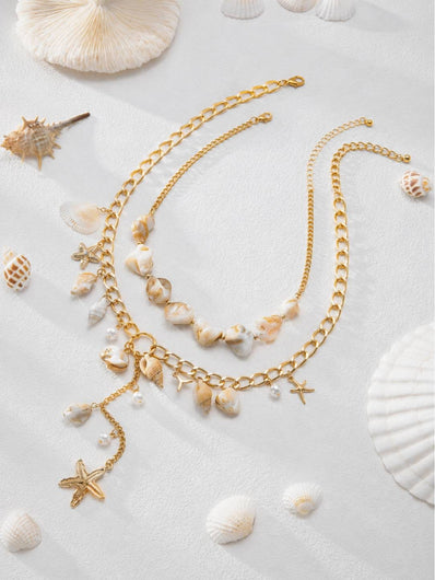 Both gold-tone shell necklaces styled together on a sandy white surface surrounded by real seashells and coral pieces